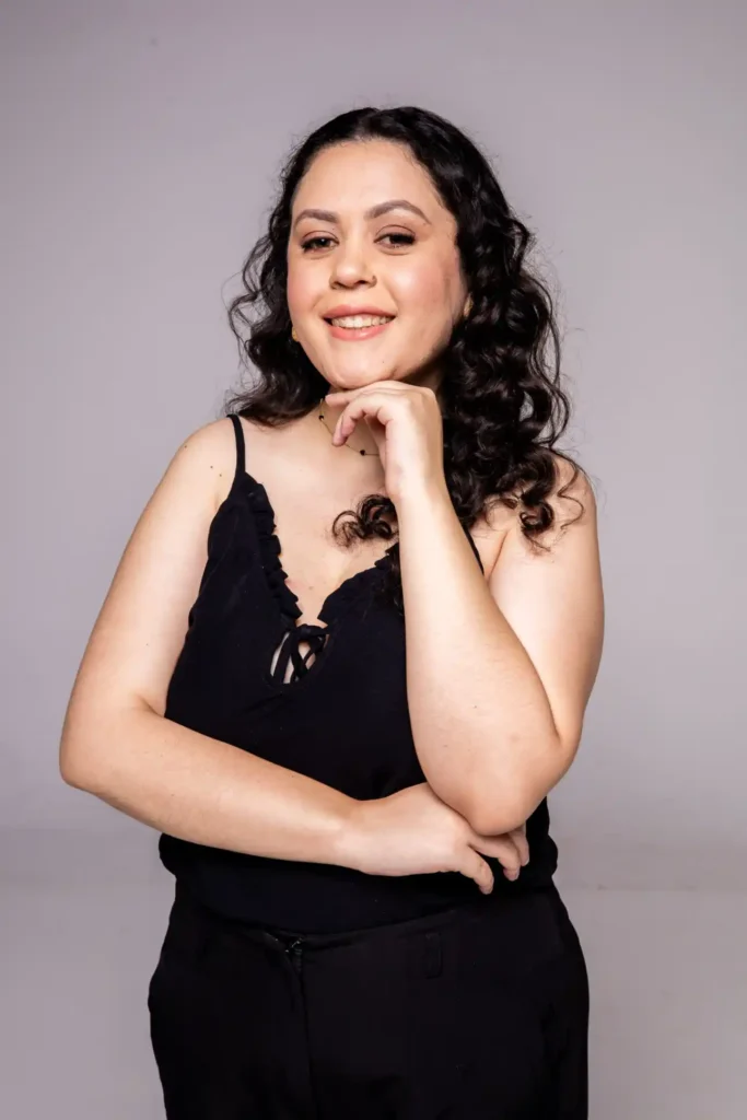 Mulher sorridente em estúdio com cabelo cacheado, usando blusa preta sem mangas e posando com a mão no queixo, fundo cinza claro.