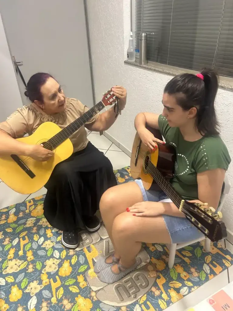 Ensaio musical em casa: duas pessoas tocam violão sentadas no chão, com uma mulher cantando ou acompanhando o ritmo e a outra segurando a guitarra acústica.