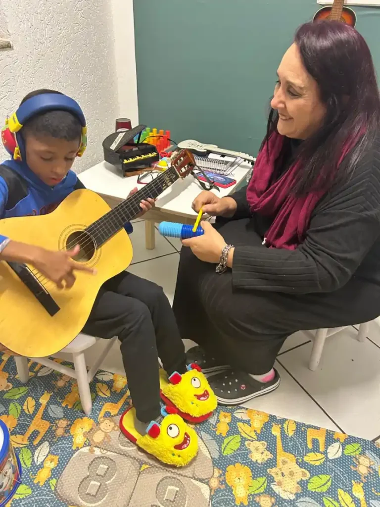 A professora e uma criança brincam e aprendem com música em uma sala, enquanto a criança toca violão e a adulta ajuda a acompanhar o ritmo com um instrumento colorido.