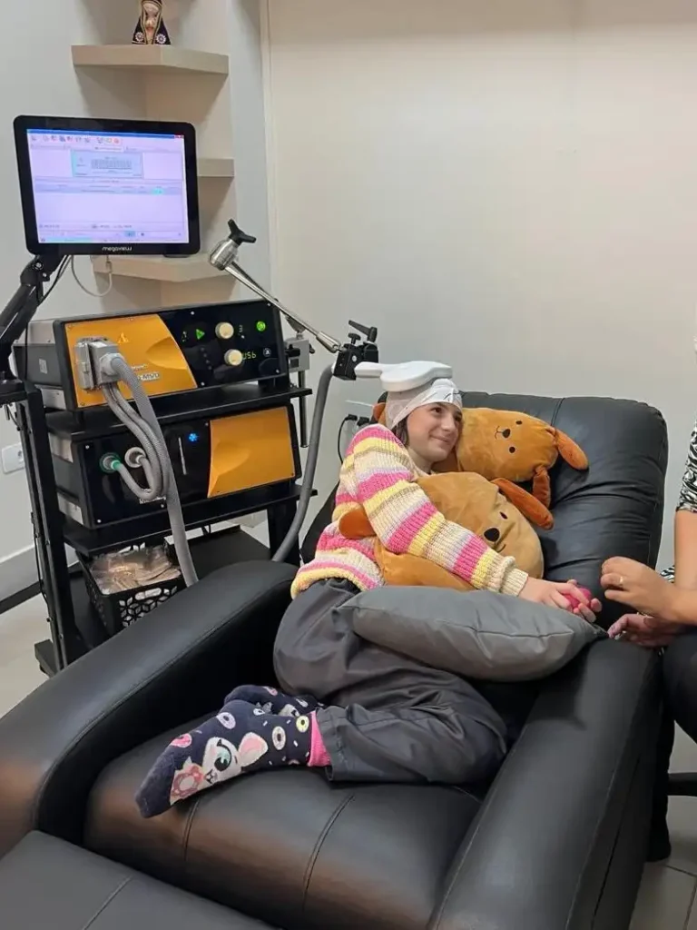 Paciente sentada em maca em clínica de fisioterapia, usando touca branca e segurando um ursinho de pelúcia, enquanto equipe utiliza equipamento de terapia com monitor ao fundo