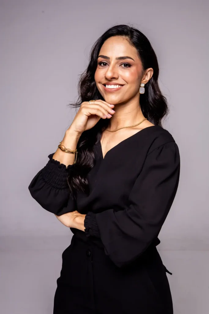 Mulher sorridente em estúdio, com cabelo longo escuro e vestido preto, usando brinco e colar dourados, posando com uma mão no queixo contra fundo claro.