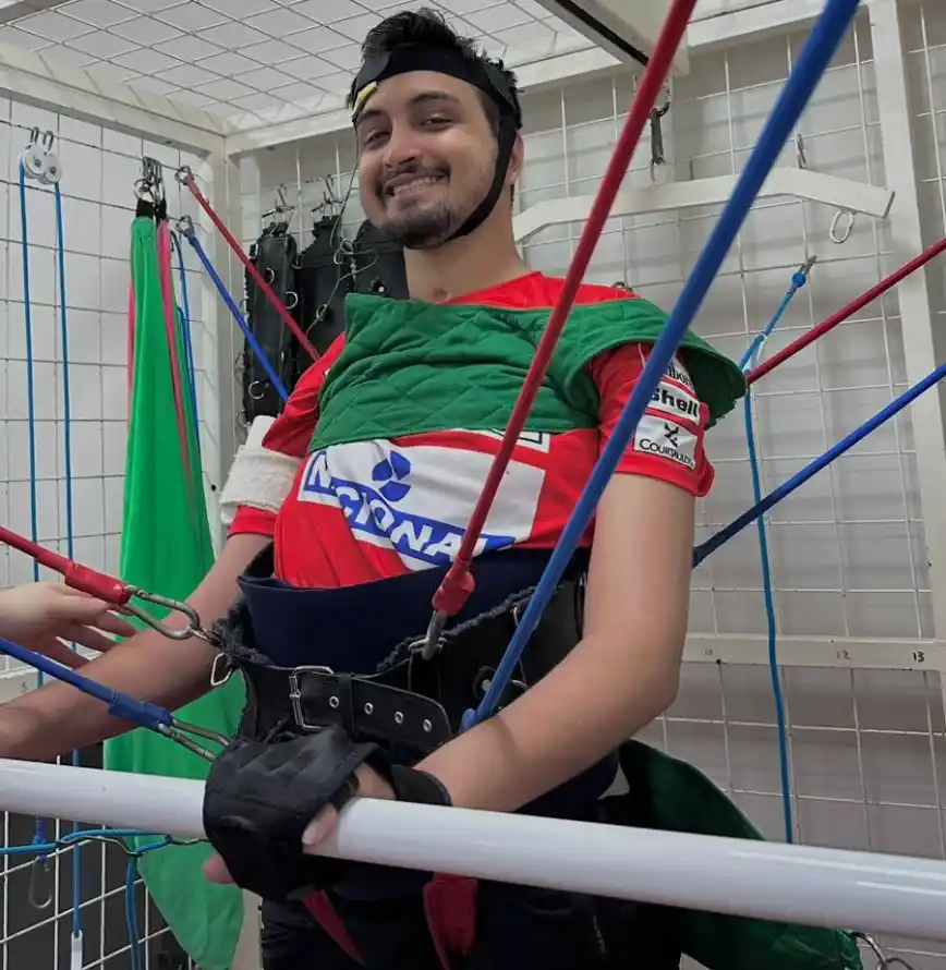 Homem usando equipamento de paraquedismo com arnês e capacete, com colete verde e vermelho, sentado em um simulador de voo dentro de uma estrutura com tubos coloridos.