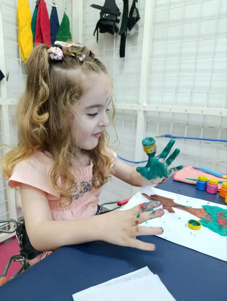 Criança em ateliê de artes pintando com a mão em uma bancada branca, com tintas coloridas ao lado, fazendo pintura durante atividade criativa.