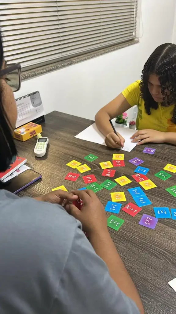 Sala de aula durante uma atividade com cartões coloridos de operações matemáticas, onde crianças e um adulto escrevem e resolvem contas em uma mesa com papel.
