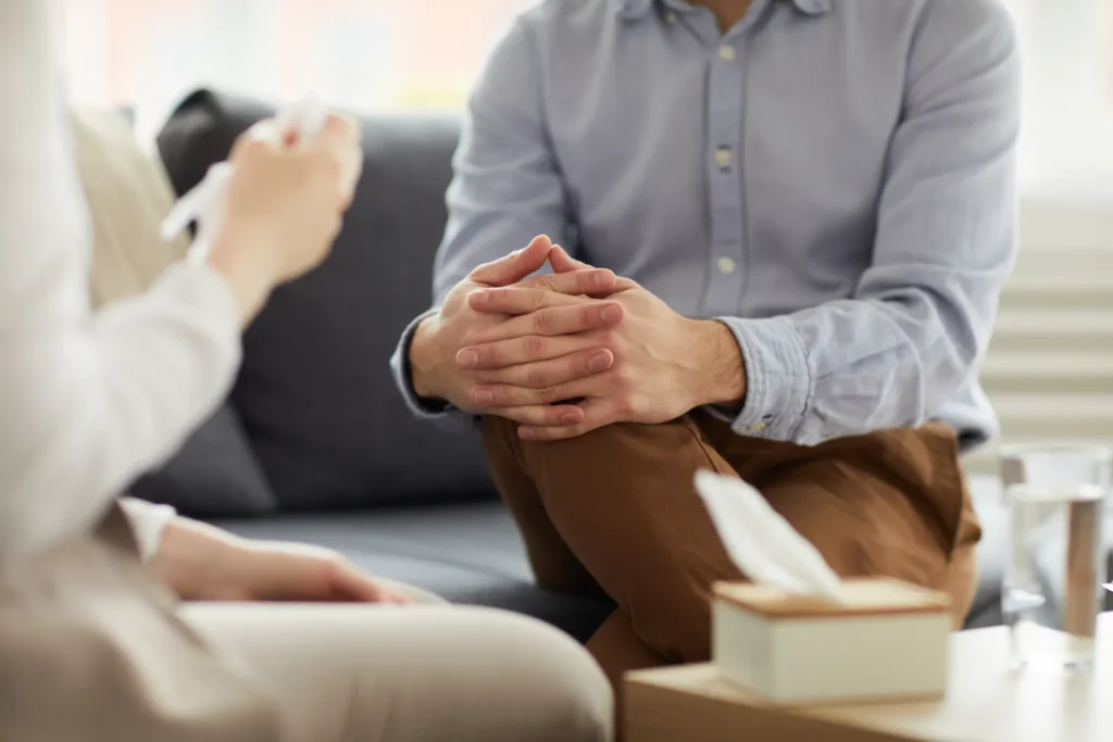 Mãos de um consultor ou terapeuta com as mãos entrelaçadas à frente, enquanto uma pessoa ao fundo recebe atendimento em ambiente acolhedor, sugerindo suporte e orientação profissional.
