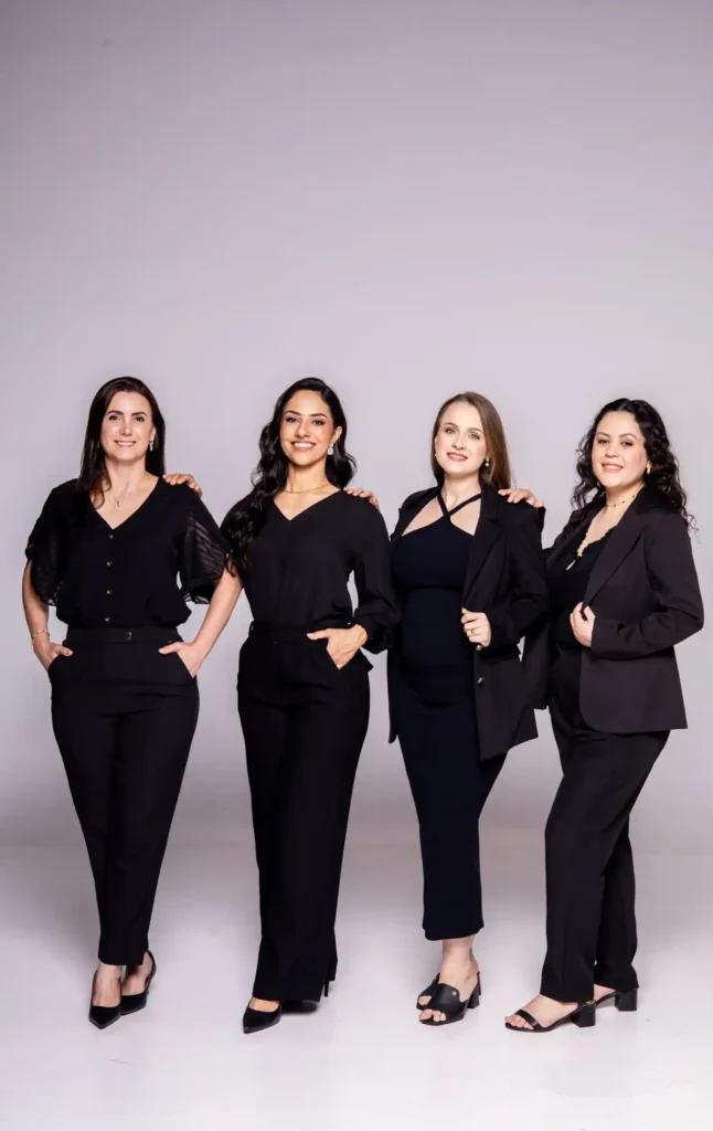 Foto de quatro mulheres vestidas com ternos e blusas pretas em estúdio, sorrindo e posando juntas contra um fundo claro, com aparência profissional.