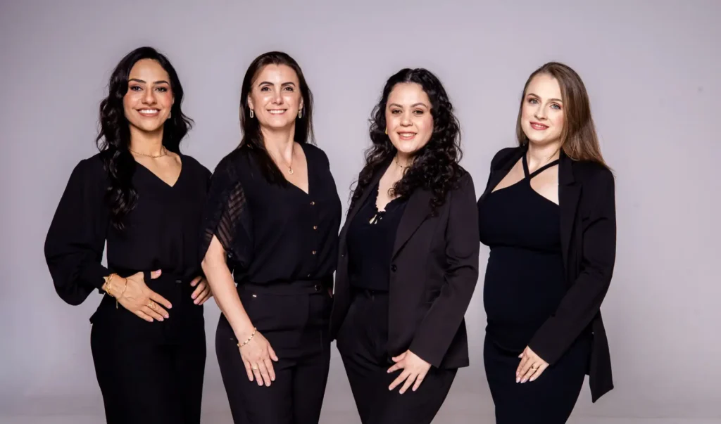 Foto de quatro mulheres em trajes sociais pretos, posando juntas em estúdio com fundo claro, com expressões sorridentes e postura profissional.