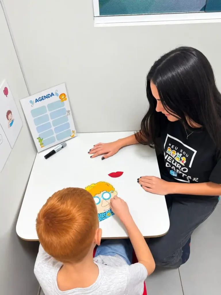Educadora acompanha uma criança enquanto preenche uma agenda infantil colorida com adesivos e canetinhas sobre uma mesa branca, em ambiente escolar.