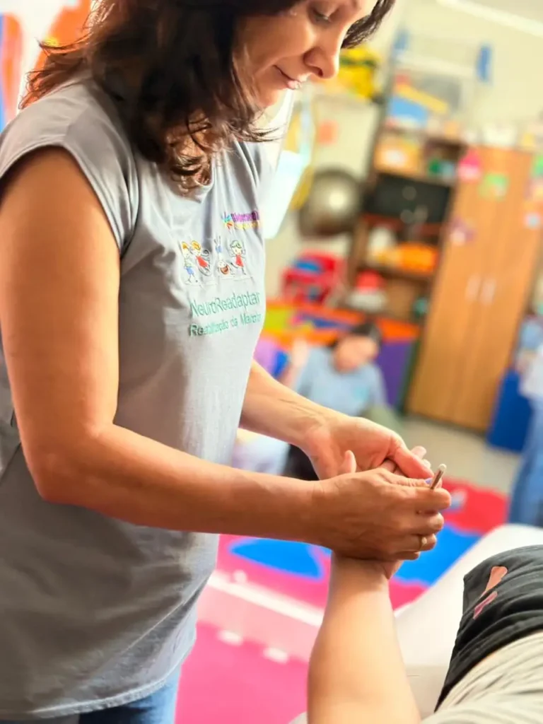Profissional realiza atendimento de fisioterapia com movimento de alongamento em pessoa sentada, em ambiente clínico com brinquedos ao fundo.