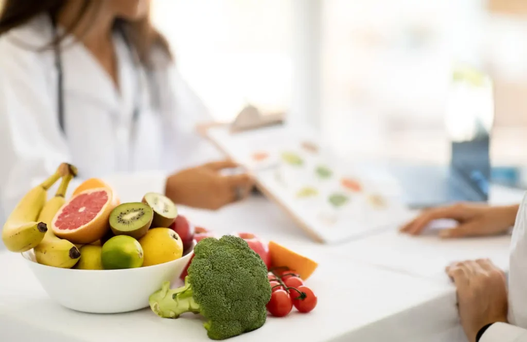 Frutas frescas em uma tigela sobre a mesa, com banana, kiwi, uvas, grapefruit e abacate; ao fundo, uma pessoa preparando um prato em cozinha clara, clima saudável.