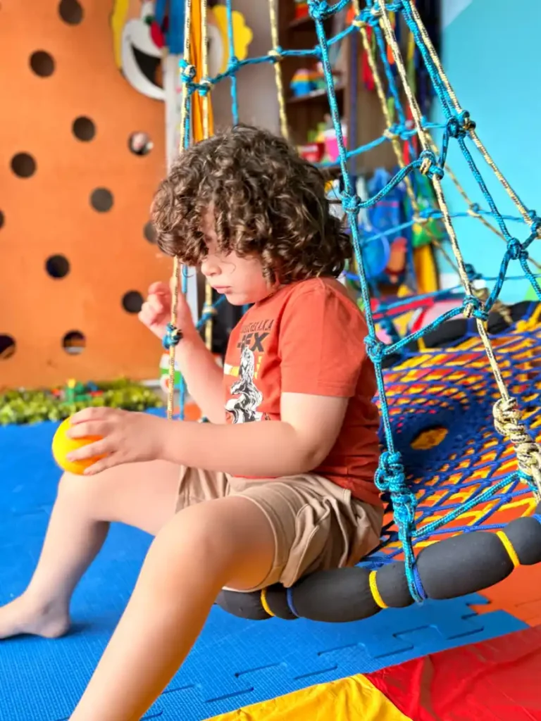 Criança com cabelo cacheado brincando em um pula-pula inflável com rede azul e amarelo, segurando uma bola, em ambiente externo colorido.