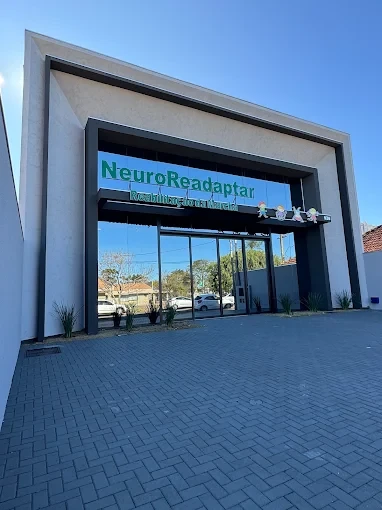 Entrada de um prédio com fachada moderna e pórtico, placa “NeuroReadaptar” com leitura clara e carros estacionados ao fundo sob céu azul, vista em ângulo baixo.