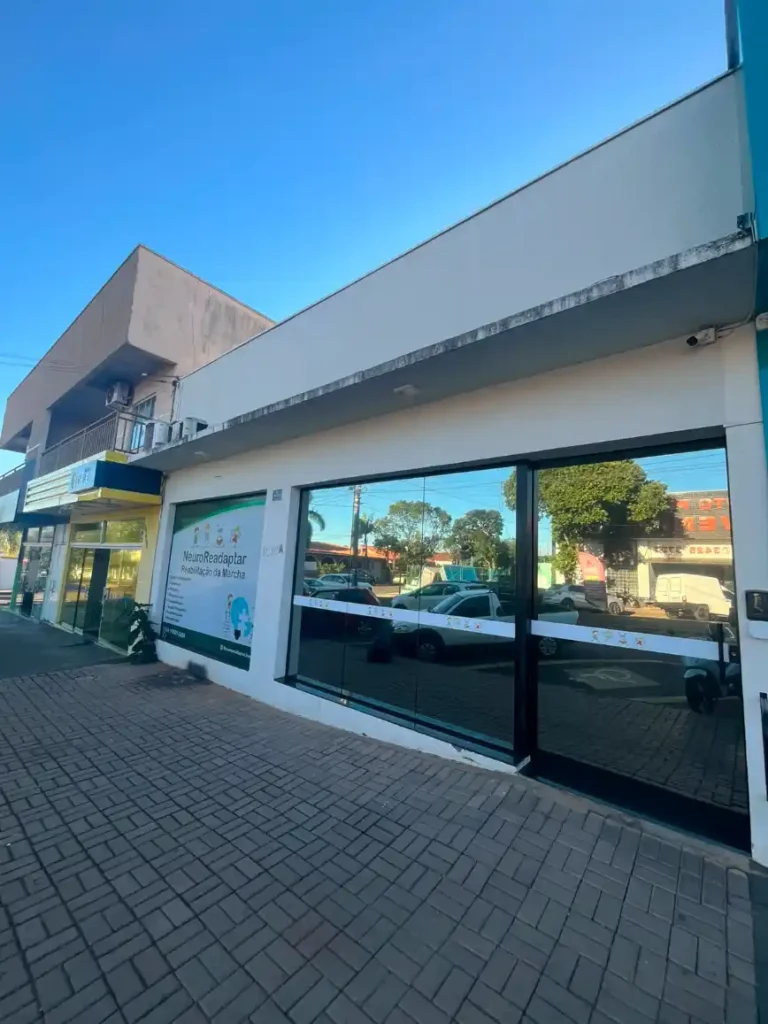 Fachada de um prédio comercial em ambiente urbano, com janelas de vidro grandes e faixa superior cinza, em céu azul. Ao lado, placa indica o serviço NeuroReadaptar.