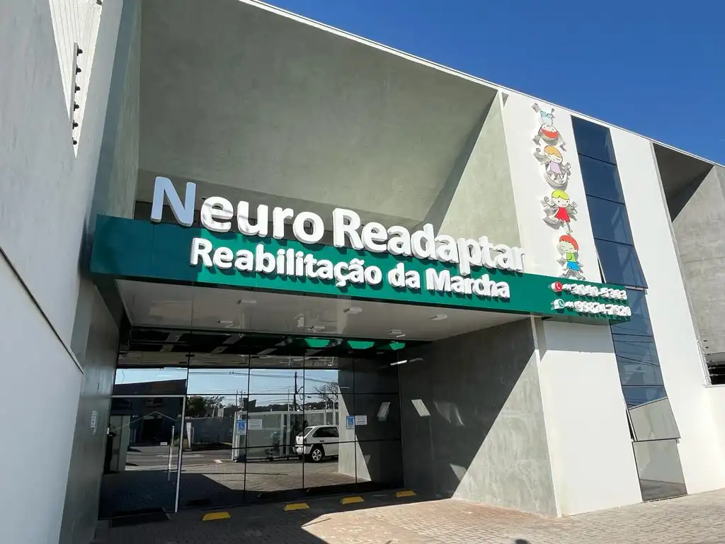 Fachada do prédio “Neuro Reagapinta” com banner “Reabilitação da Maria” e ilustrações de personagens na entrada, sob céu azul. A construção moderna tem vidro e estrutura geométrica.